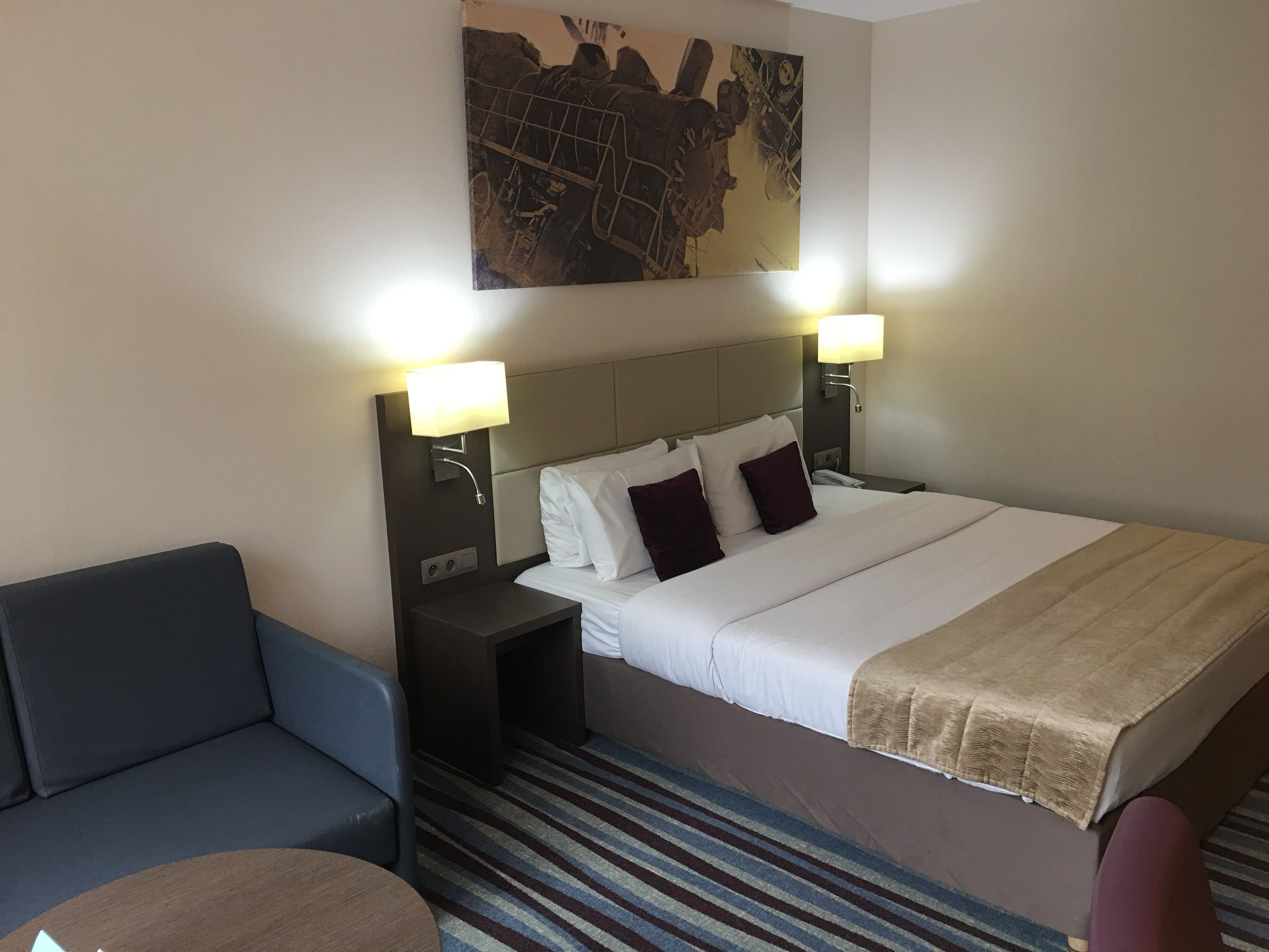 A hotel room with a large bed, two bedside lamps, a headboard, a small table, and a blue armchair.