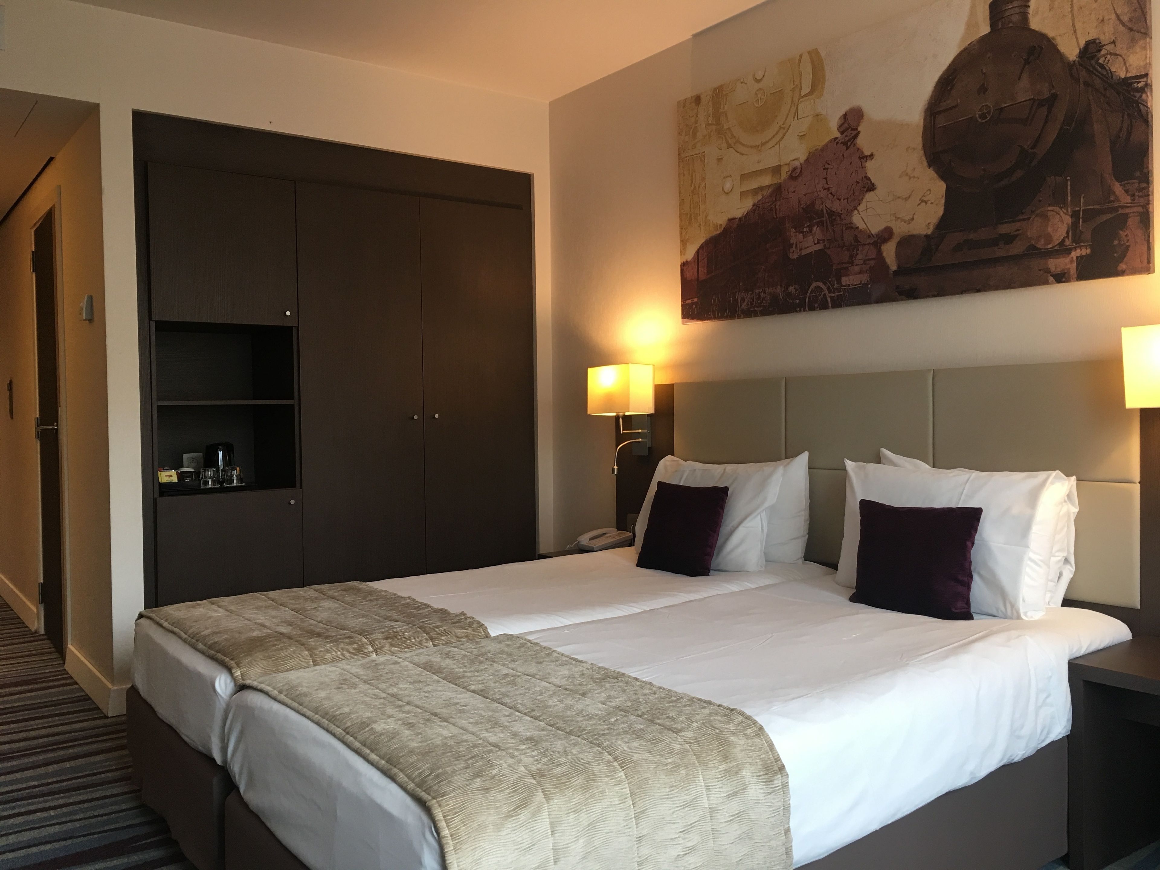 A hotel room with a large bed, a wardrobe, a painting on the wall, and a bedside lamp.