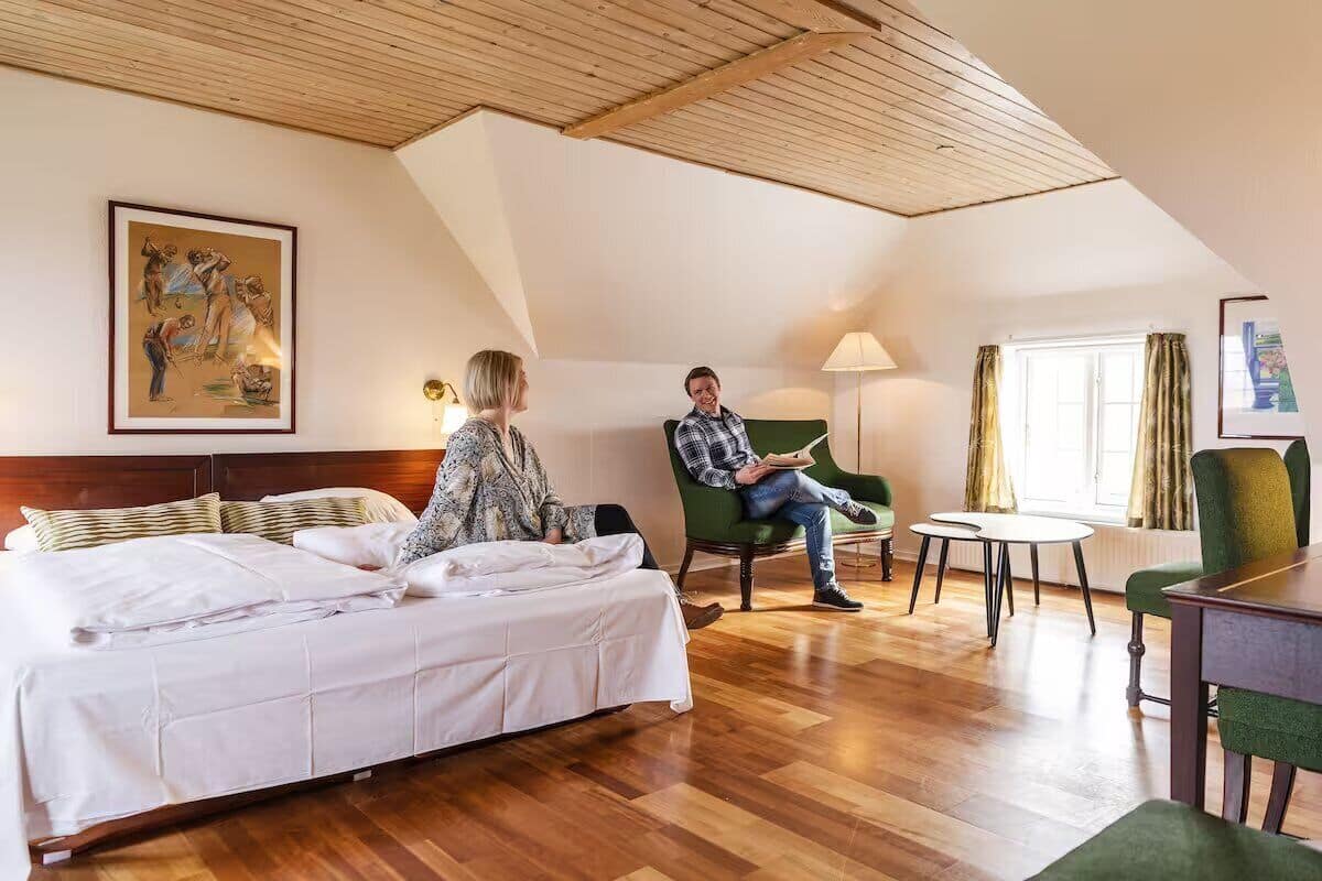 A hotel room with a bed, a green armchair, a small table, and a window with curtains.