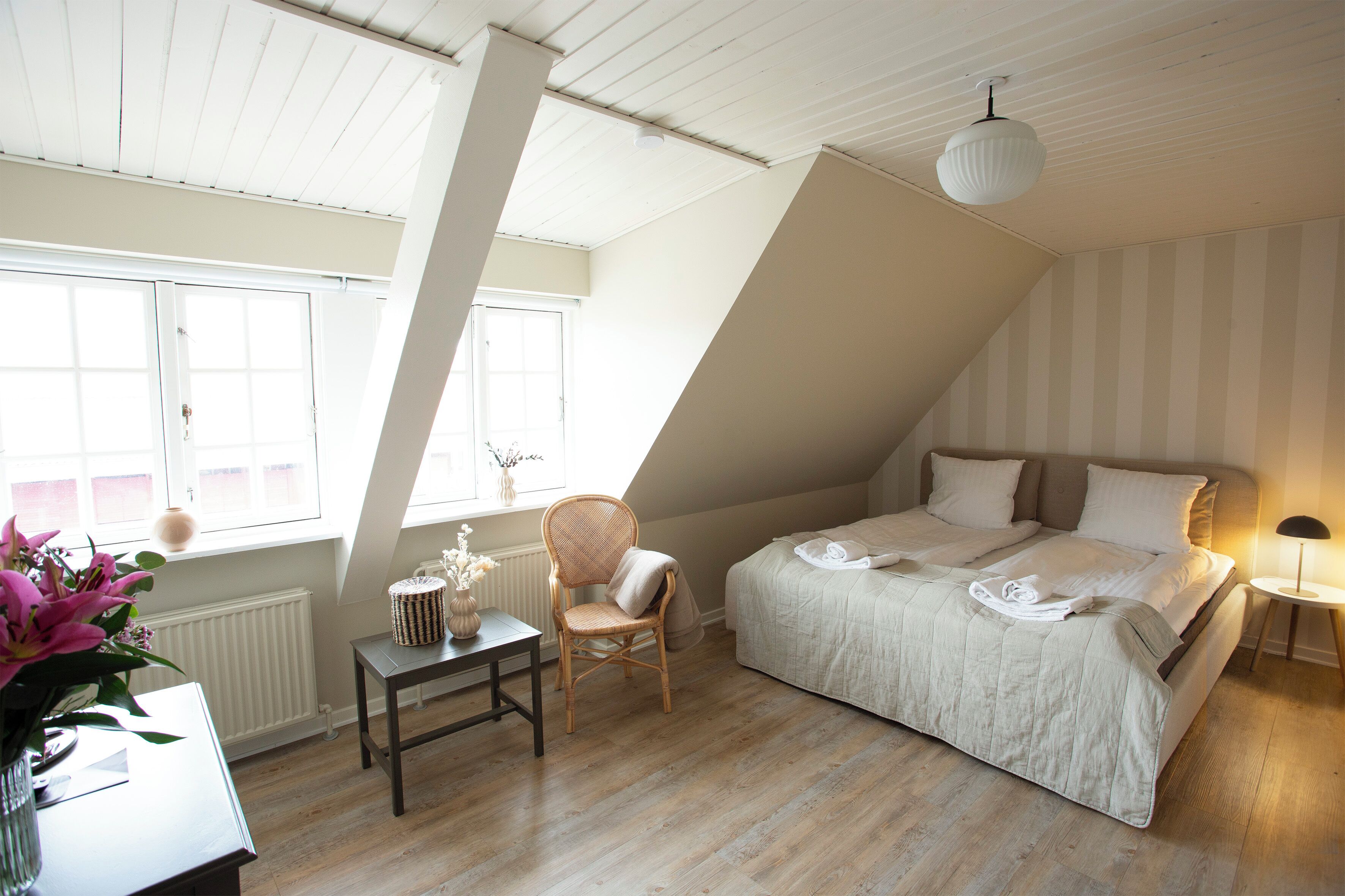 A bedroom with a sloped ceiling, a bed with white linens, a wicker chair, a small table with a vase of flowers, and a bedside lamp.
