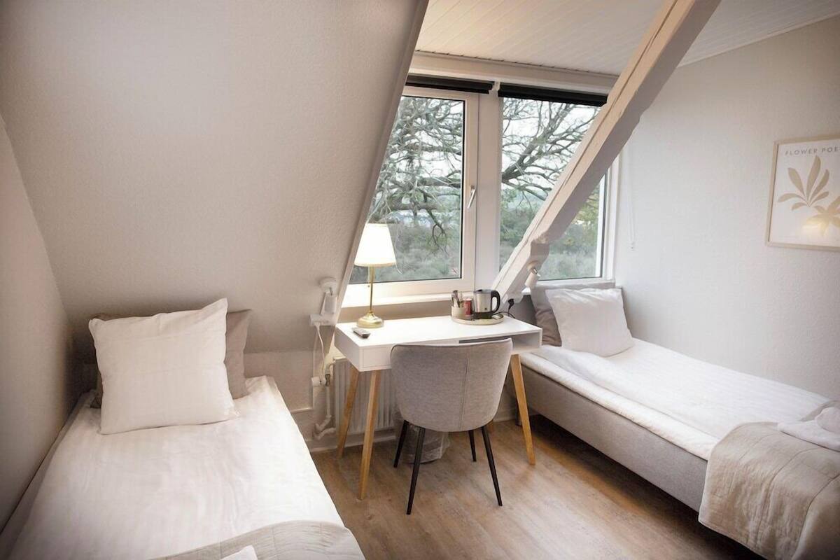 A small, well-lit room with a bed, a desk, and a chair, a window with a view of trees, and a framed picture on the wall.