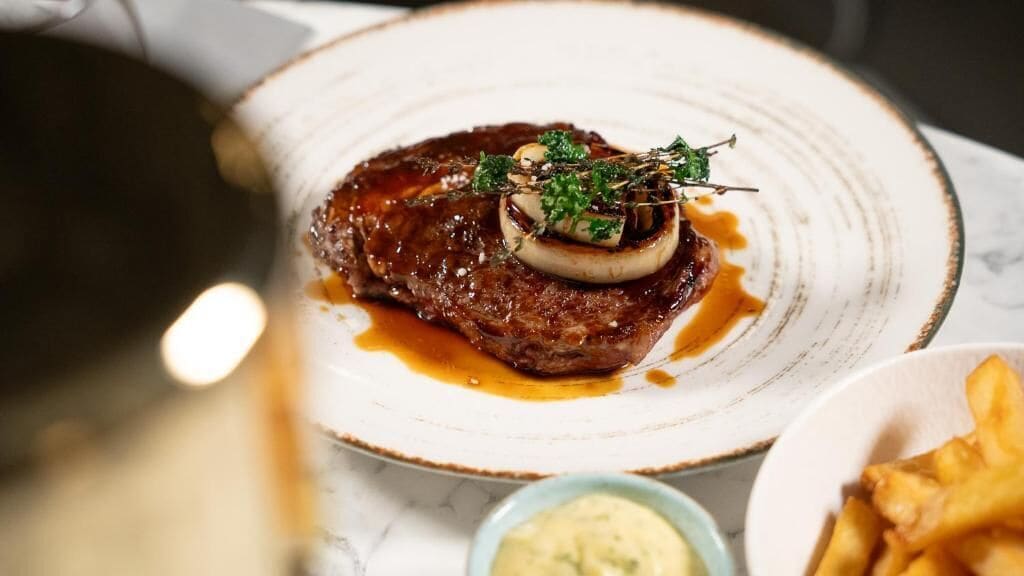 A steak with a sauce and garnish on a plate, accompanied by fries and a small bowl of sauce.