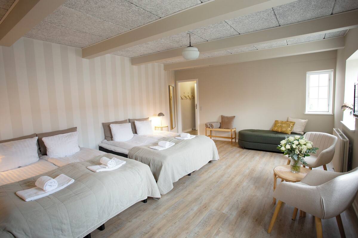 A hotel room with two beds, a sofa, a chair, a small table with a vase, and a window.