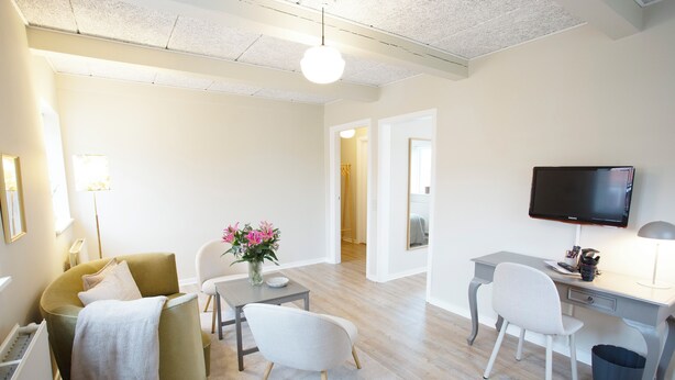 A modern living room with a sofa, chairs, a desk, a TV, and a vase with flowers. - HotelVFjorden (Lemvig)