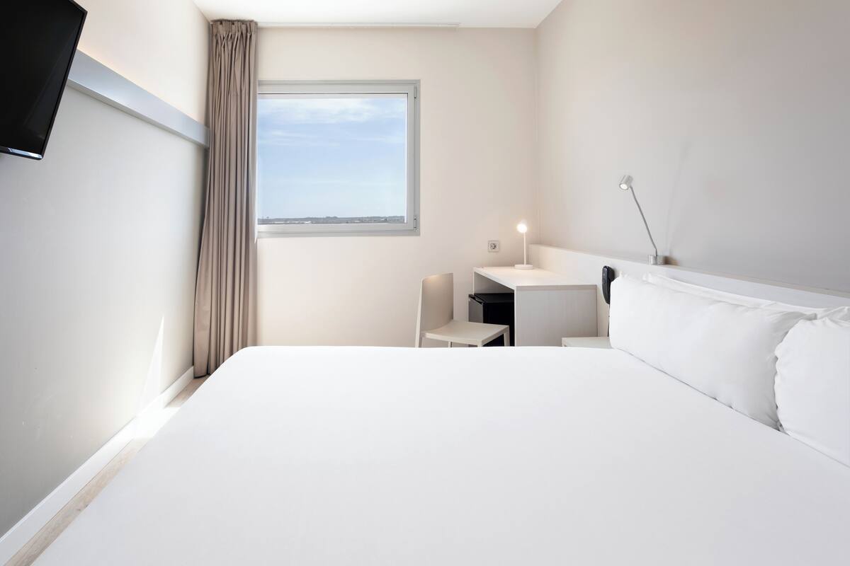 Una habitación de hotel con una cama grande, un escritorio, una silla, un televisor y una ventana con cortinas.