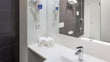 A modern bathroom with a white sink, two hair dryers mounted on the wall, and a shower area visible in the mirror reflection.