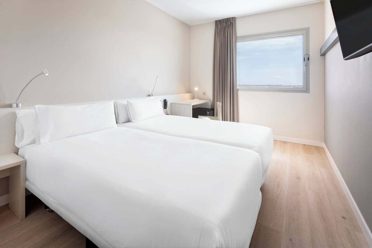 Una habitación de hotel moderna con dos camas, un escritorio, una silla, una lámpara y una ventana con cortinas.