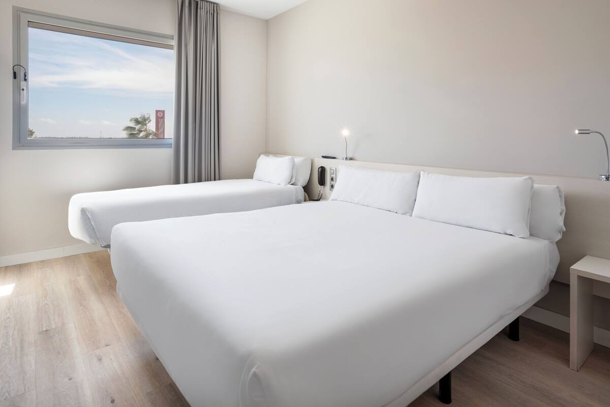 Habitación de hotel con dos camas individuales, una mesita de noche, una lámpara, un teléfono y una ventana con vista a palmeras.