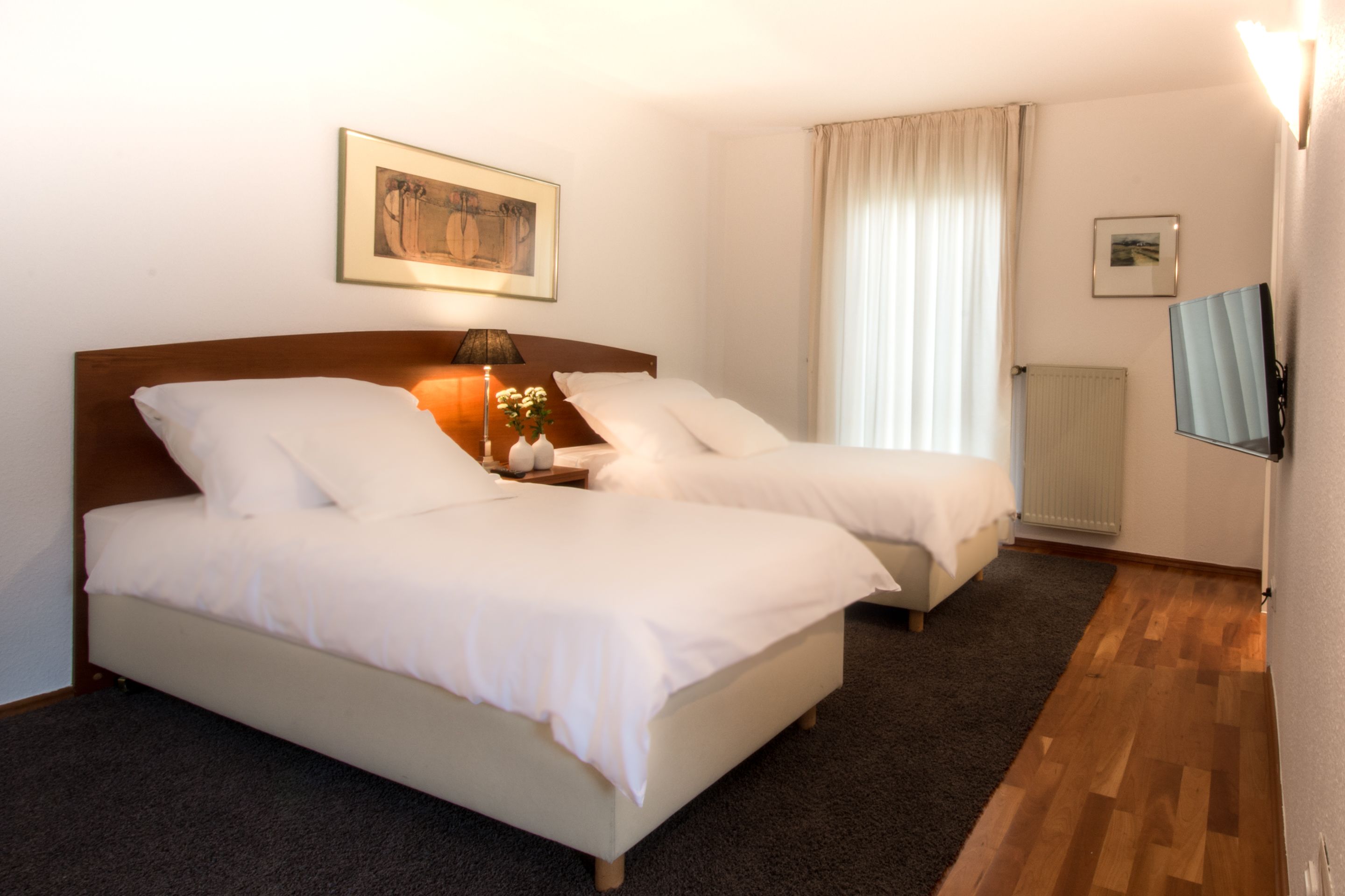 Ett hotellrum med två separata sängar, en sänggavel i trä, ett nattduksbord med en lampa, en inramad tavla på väggen, ett fönster med gardiner och ett element.
