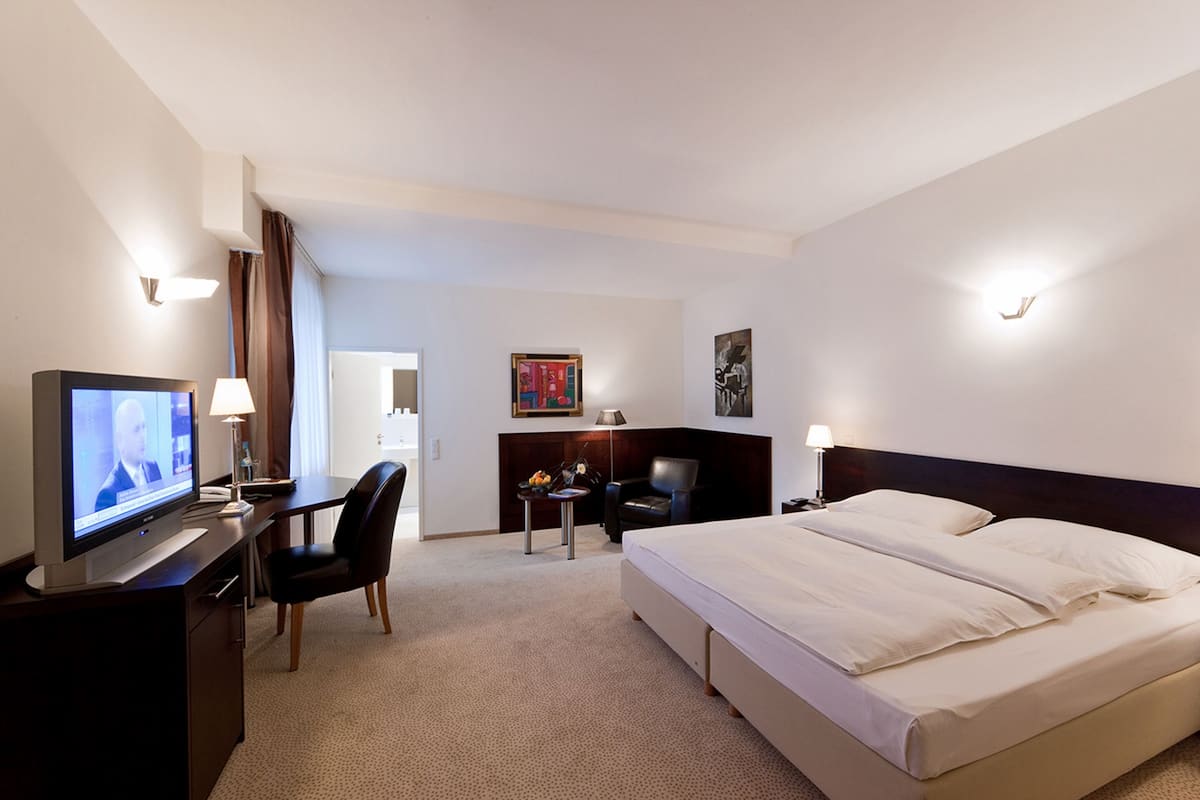 Habitación de hotel con cama, escritorio con televisor, silla, mesita con lámpara y un cuadro en la pared.