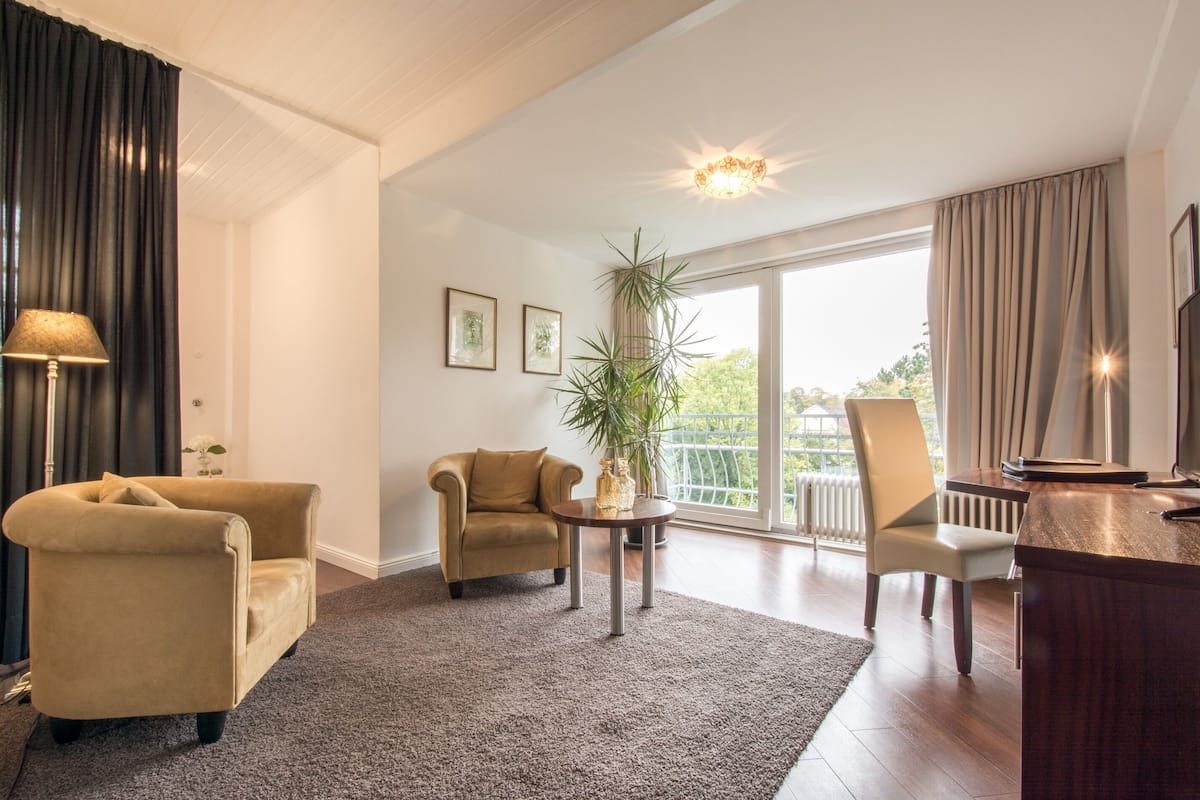 Una sala moderna con un sofá, un sillón y una mesa de centro. Hay un ventanal con cortinas, una planta y vistas al exterior.