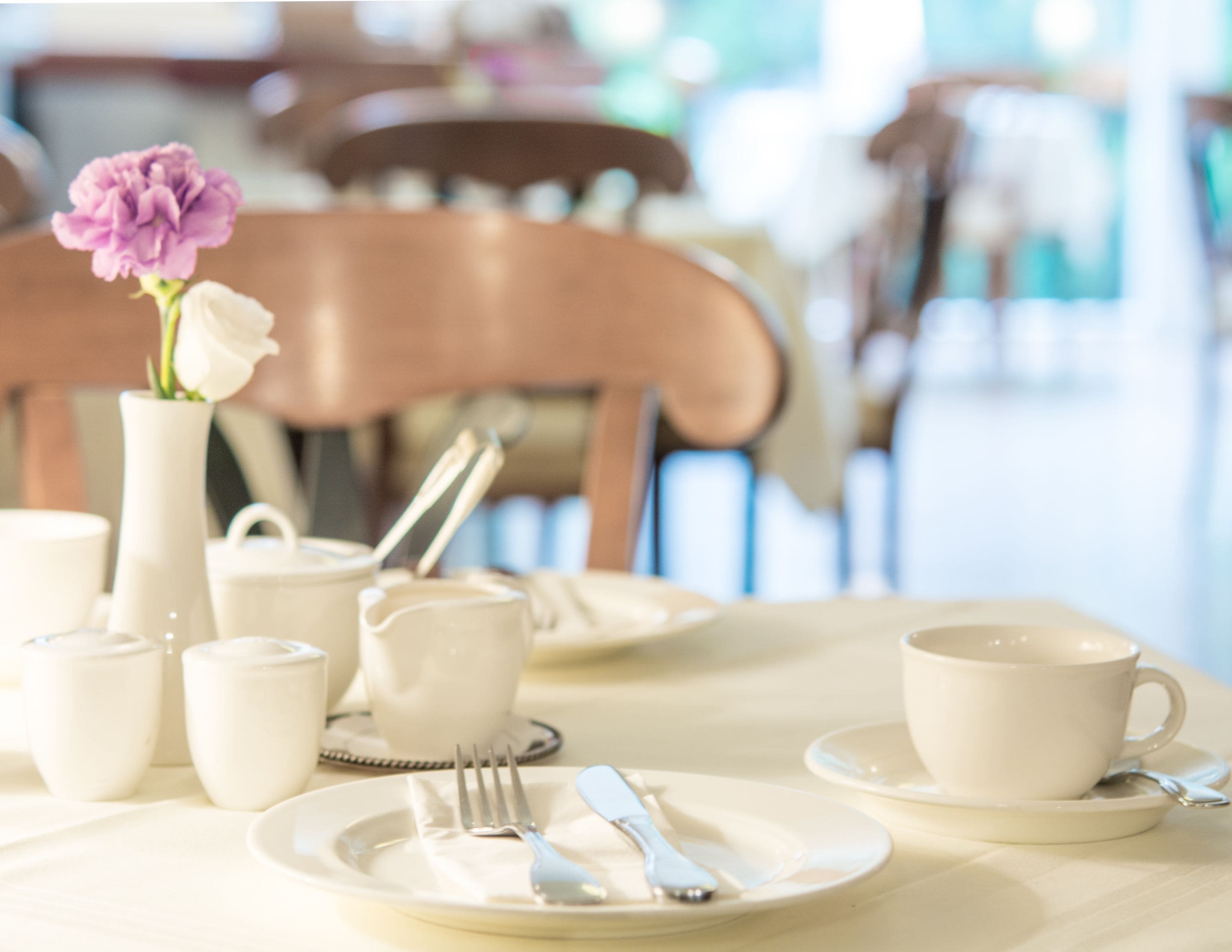 Una mesa de comedor dispuesta con platos blancos, tazas y cubiertos, un florero con una flor morada y una jarra blanca.