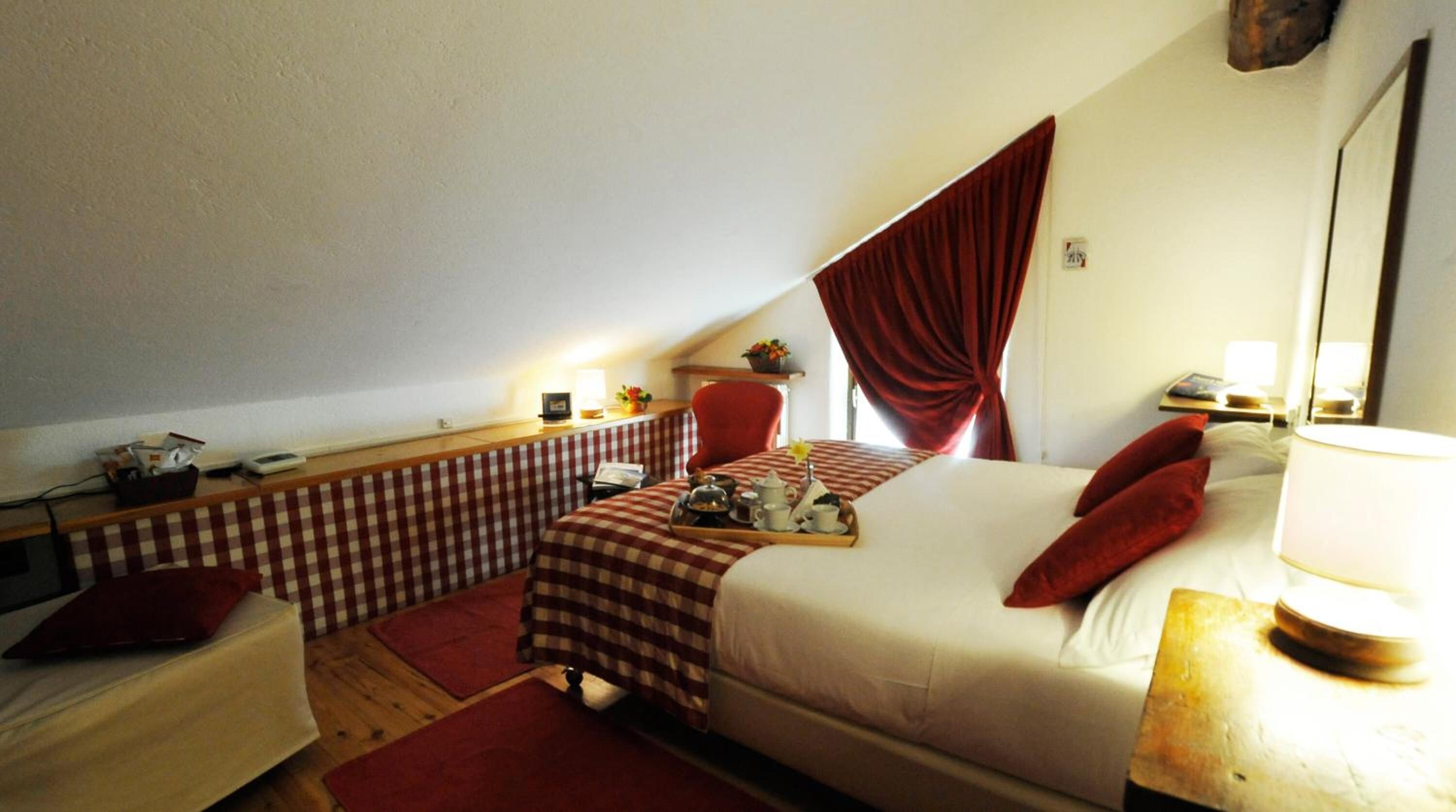 Una camera d'albergo accogliente con una panca a scacchi, un letto con cuscini rossi, una sedia rossa e un tavolino con un vassoio di tè e pasticcini.