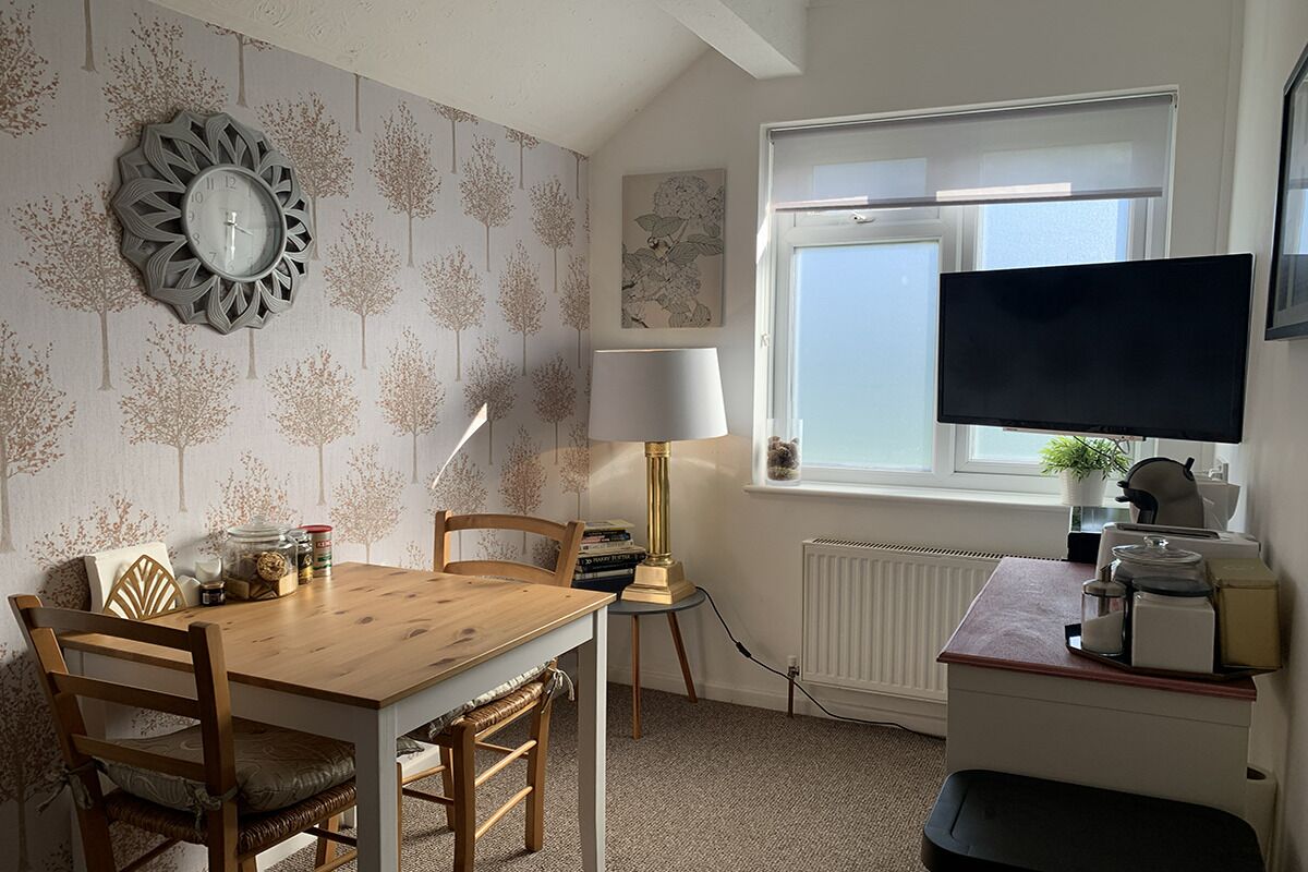 A room with a wooden dining table, chairs, a wall clock, a lamp, a television, and a window with a view.