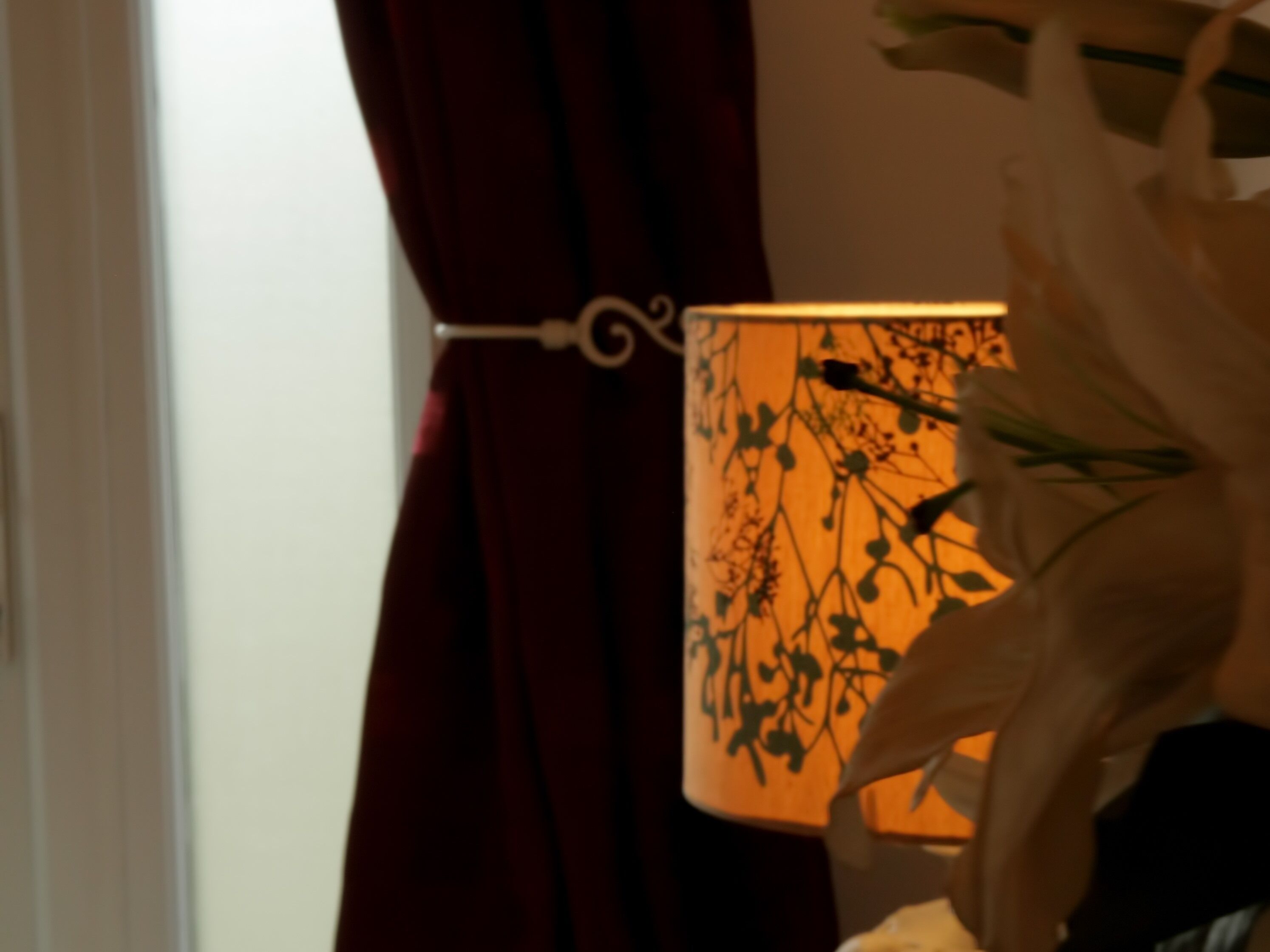 A room with a lamp on a table, a curtain, and a wall with a floral pattern.