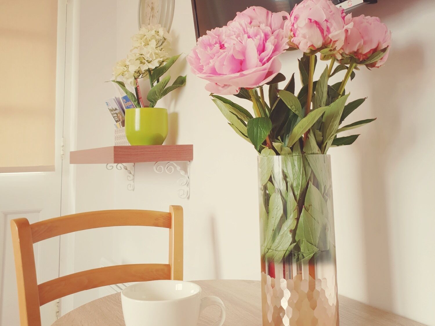A table with a vase of flowers, a green cup, and a white mug.