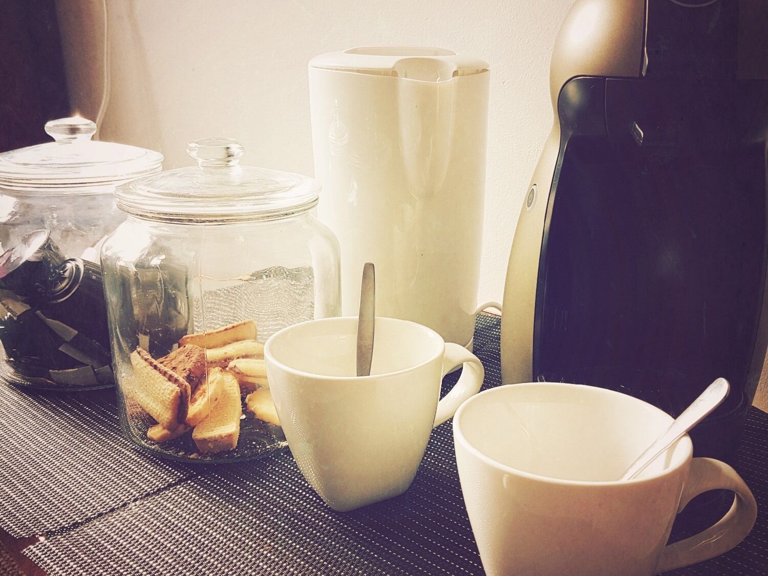 Una preparación para el desayuno con una cafetera, dos tazas, un vaso y un tarro de comida.