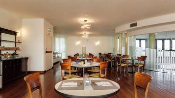 Un comedor de hotel con varias mesas redondas, sillas de madera y una cocina visible a la izquierda.