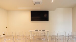 A conference room with a white table, clear plastic chairs, a flat-screen TV mounted on the wall, and a ceiling-mounted air conditioning unit. - HC Hotel Cremona Pilgrim's (Cicognolo)