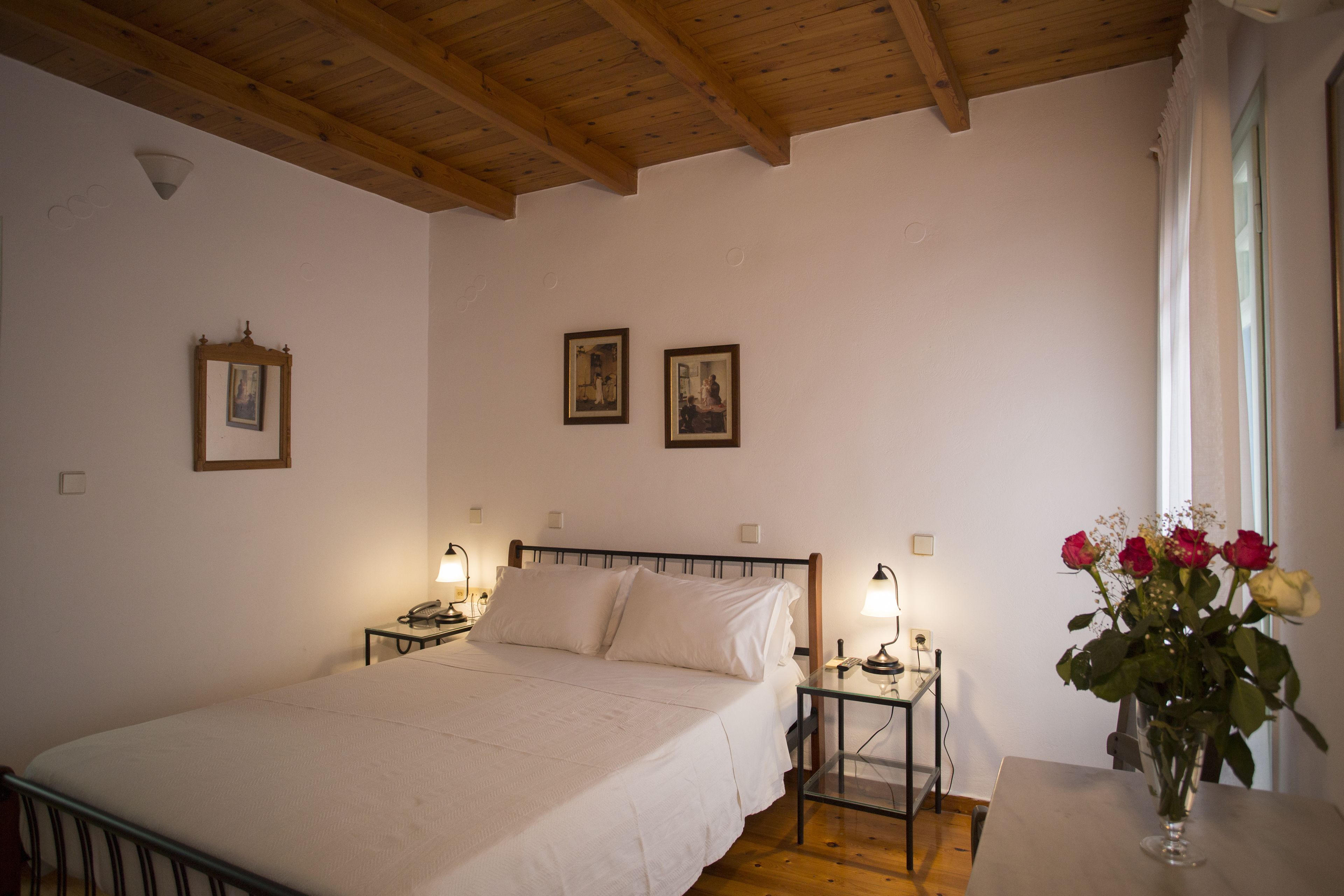 A bedroom with a wooden ceiling, a bed with white linens, bedside tables with lamps, and a vase with flowers on a table.