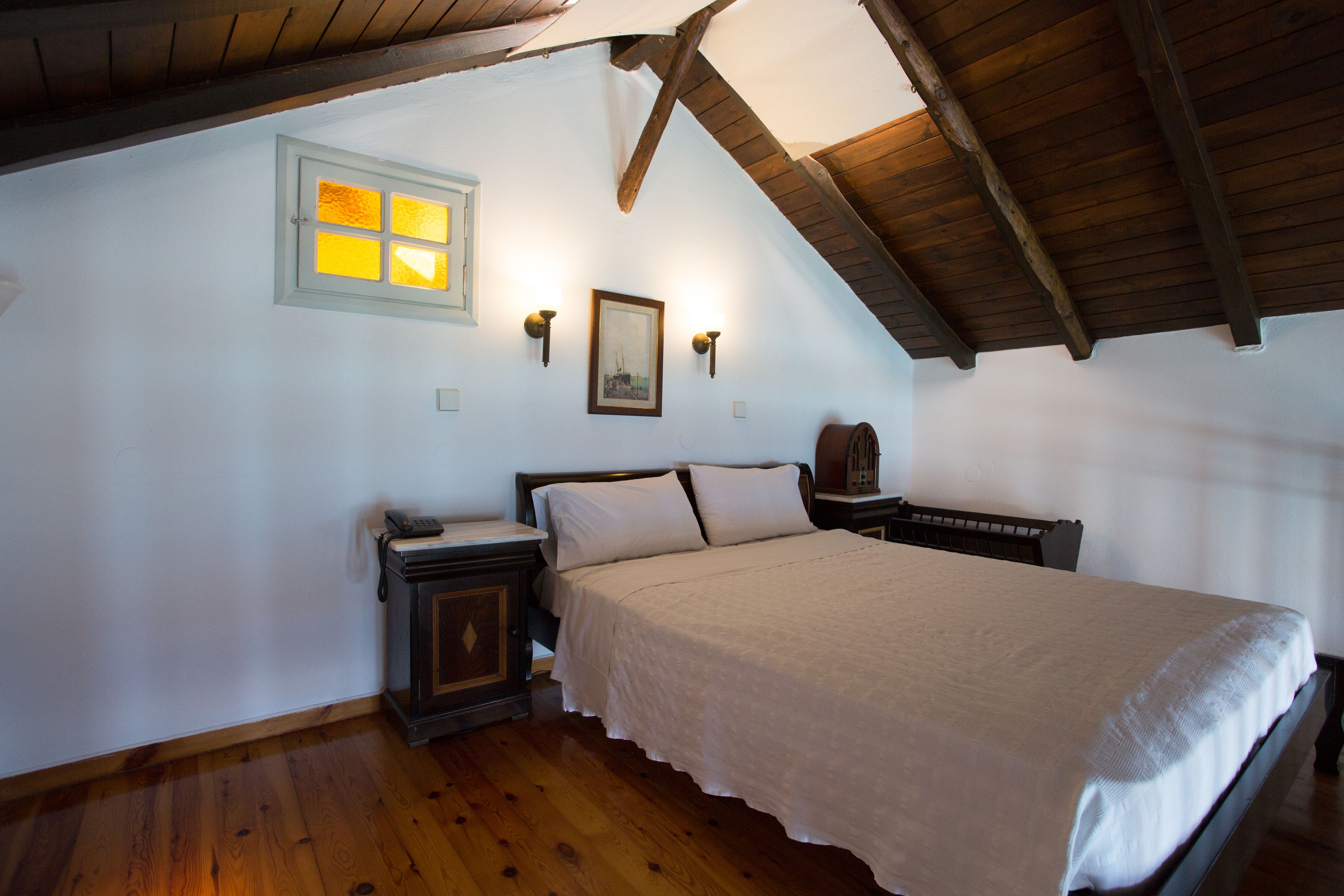 A bedroom with a wooden ceiling, a bed with a white bedspread, a nightstand, a wall-mounted lamp, and a framed picture on the wall.