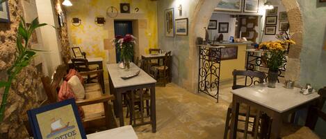 Un restaurante acogedor con paredes de piedra rústica, mesas y sillas de madera. Hay una pequeña mesa de exhibición con un letrero, y un jarrón con flores sobre una de las mesas.