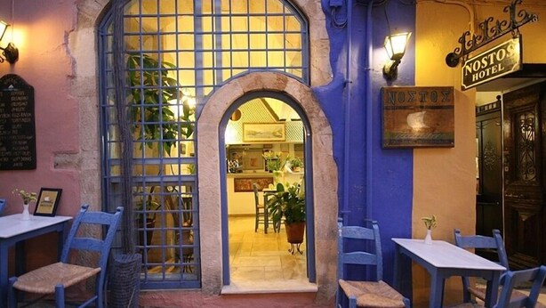 Un área de estar al aire libre con sillas y mesas azules, una amplia entrada arqueada y un cartel para el Hotel Nostos.