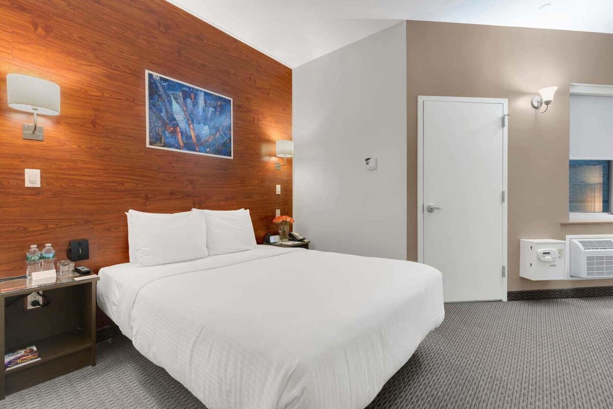 A hotel room with a bed, a nightstand, a wall-mounted lamp, a painting, a door, and an air conditioning unit.