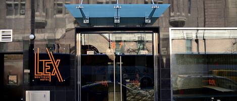 A hotel entrance with a glass door, a sign reading "LEX Boutique Hotel", and a reflection of a car and street in the glass.