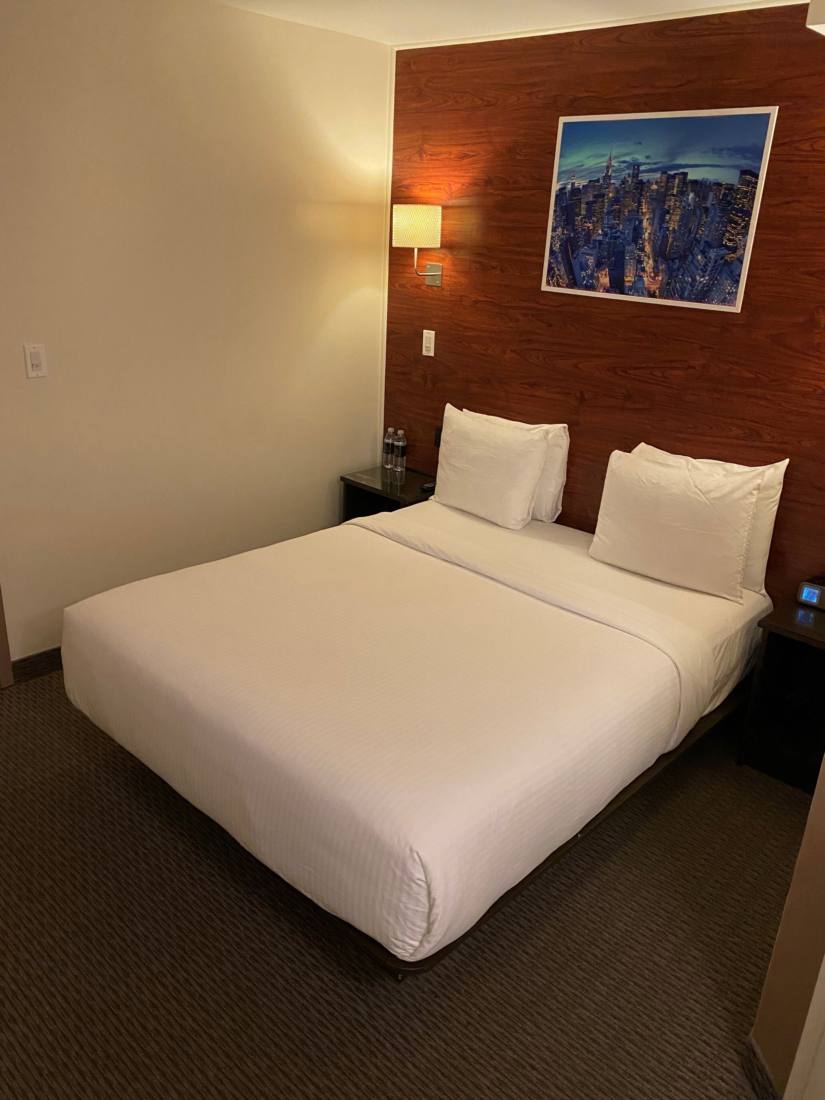 A hotel room with a large bed, a nightstand with a lamp, and a cityscape painting on the wall.