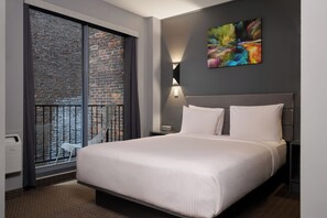 Un hotel moderno con una cama grande, vistas a un muro de ladrillo, un balcón con una silla y un cuadro abstracto y colorido en la pared.