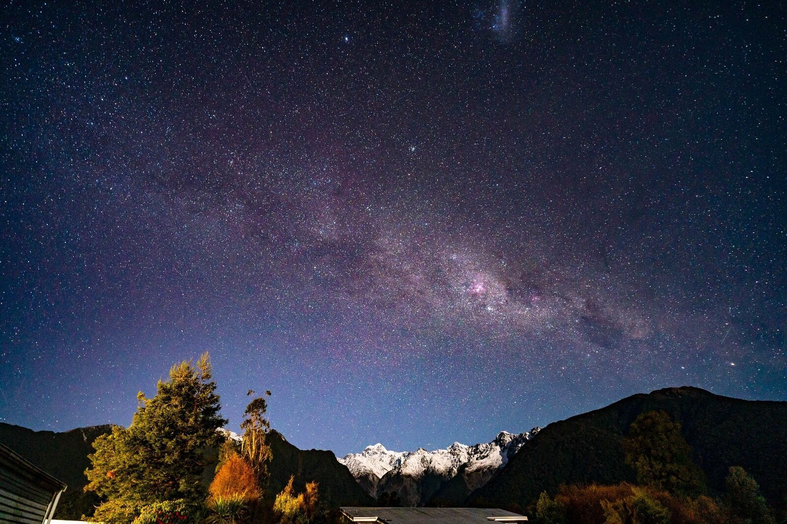 A night sky filled with stars and the Milky Way over a mountainous landscape with trees and a building.