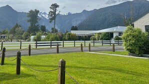 A grassy area with a white fence and chain-link barrier, a building with a sloped roof, and a mountainous backdrop.