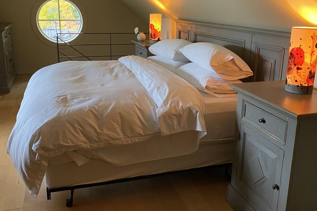 Quarto com cama, mesas de cabeceira, luminária e janela com vista para árvores.