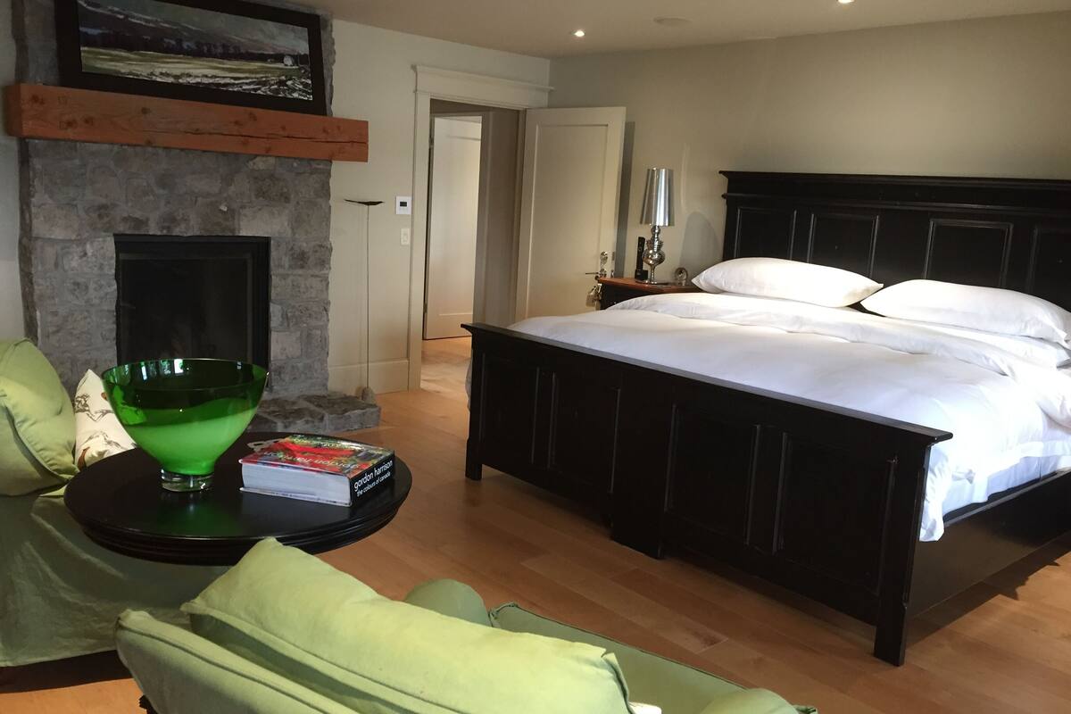 Um quarto com uma cama grande, uma lareira, uma tigela verde sobre uma mesa lateral e um quadro acima da lareira.
