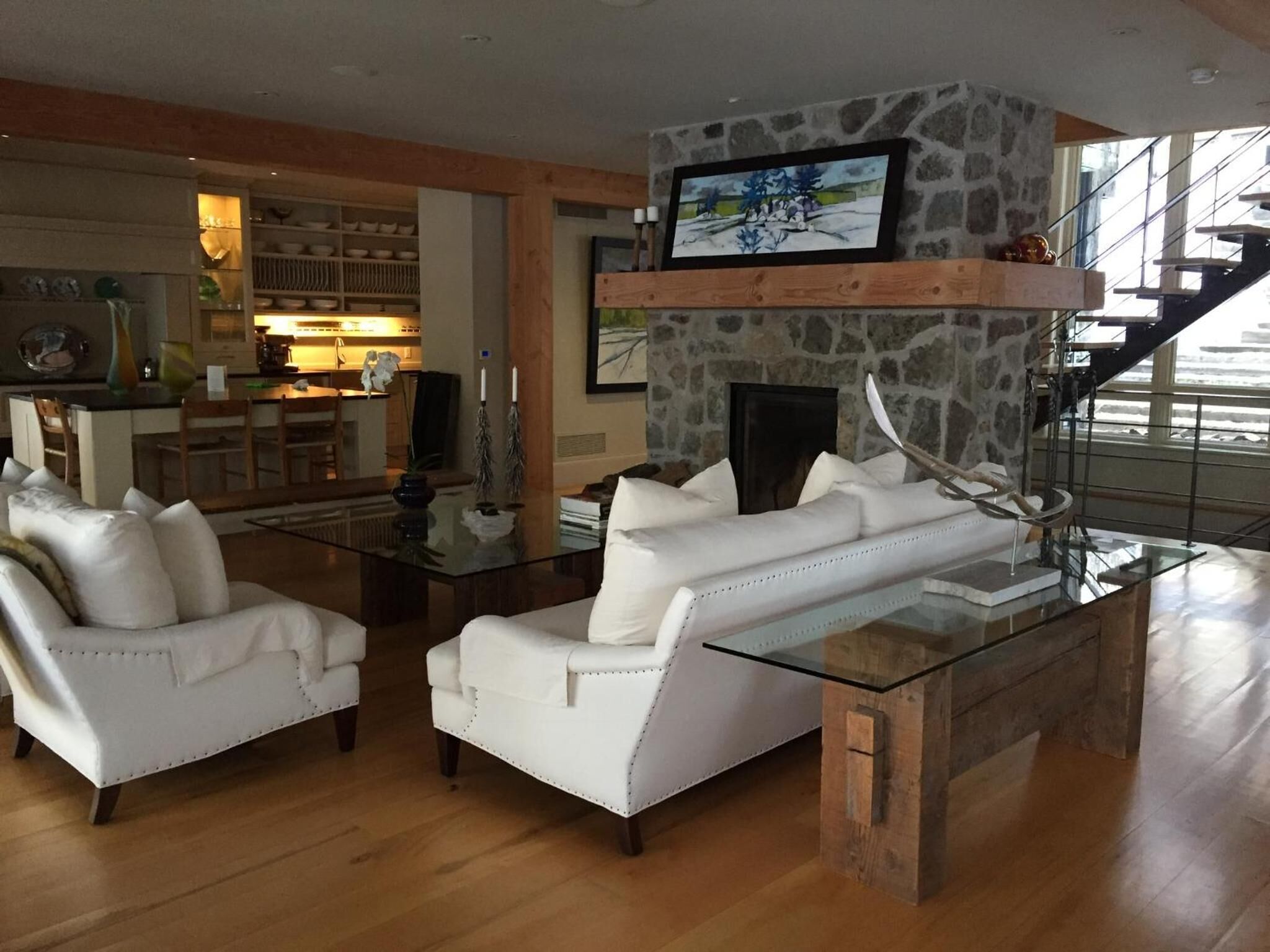 A modern living room with a glass coffee table, white leather sofas, a stone fireplace, and a kitchen in the background.