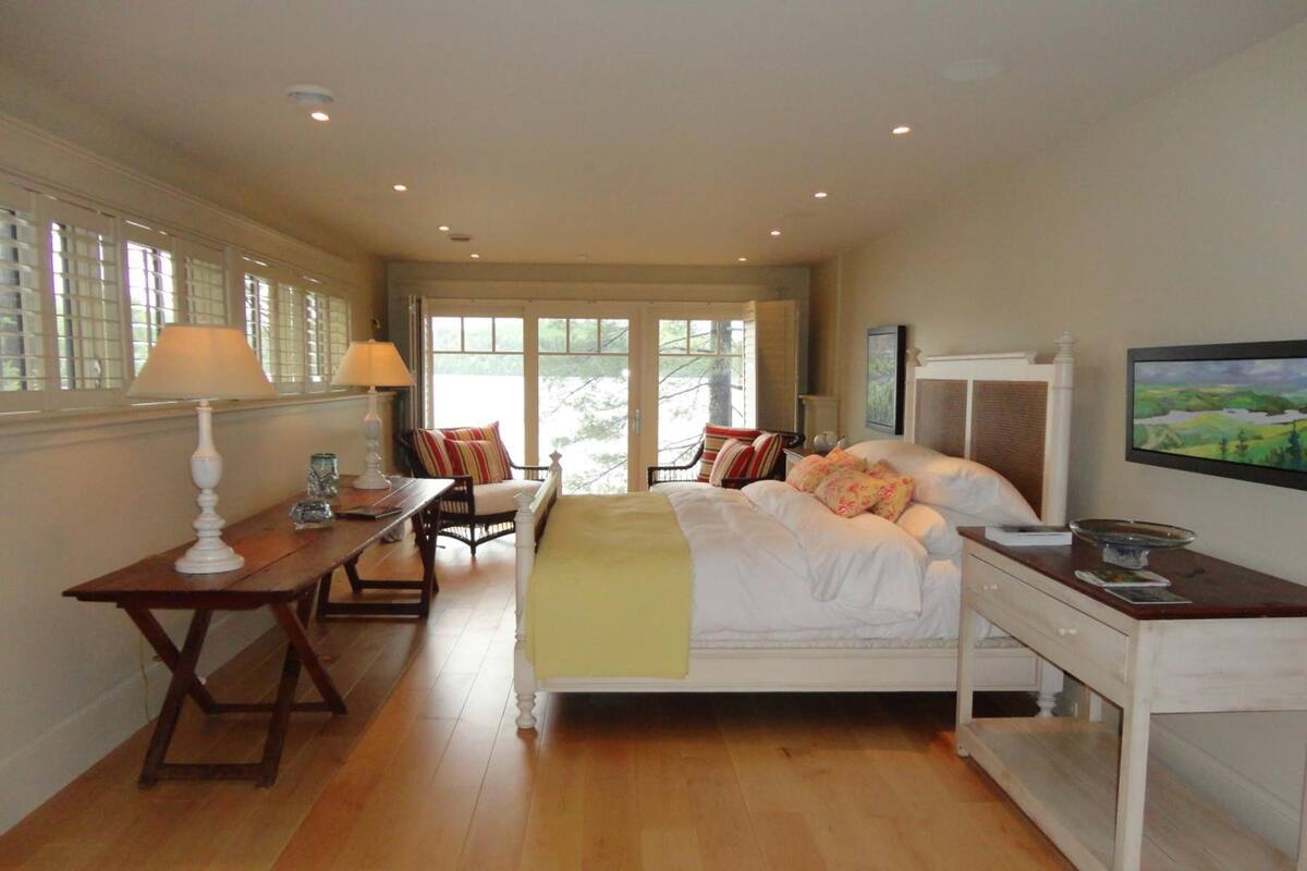 Quarto com cama grande, escrivaninha e cadeira. Tem janela com persianas e um quadro na parede.