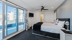 A modern bedroom with a large bed, a ceiling fan, a TV, and a view of the city through floor-to-ceiling windows. - The Pavilions Beachfront Apartments (Tauranga)