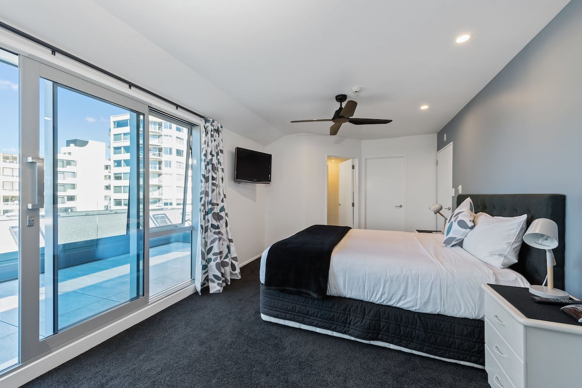 A modern bedroom with a large bed, a ceiling fan, a TV, and a view of the city through floor-to-ceiling windows.