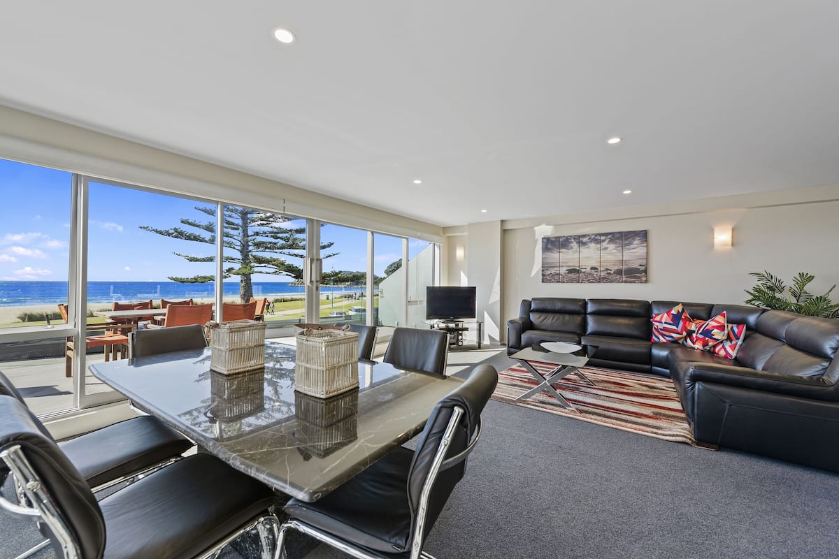 A modern living room with a large glass table, leather sofa, and a view of the beach.
