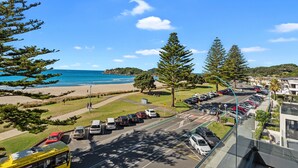 A coastal road with parked cars, a bus, and a view of the beach and ocean. - The Pavilions Beachfront Apartments (Tauranga)