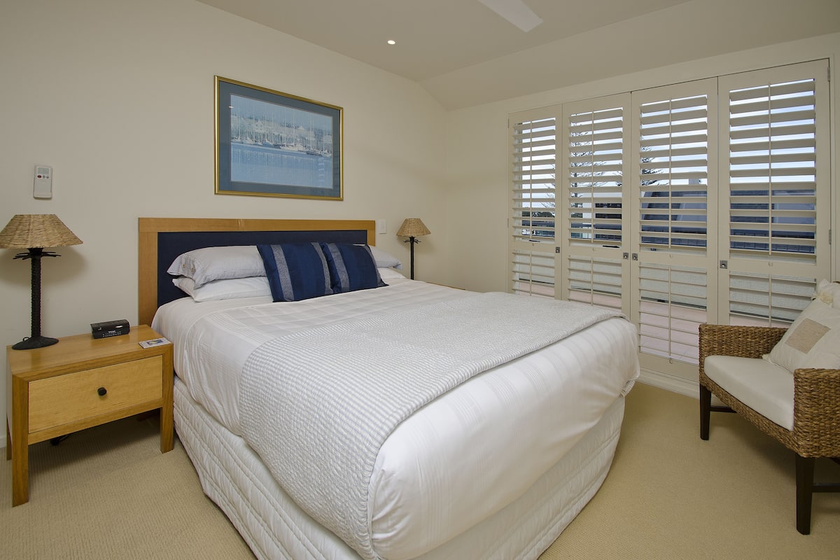 A bedroom with a large bed, two bedside tables with lamps, a wicker chair, and a window with blinds.