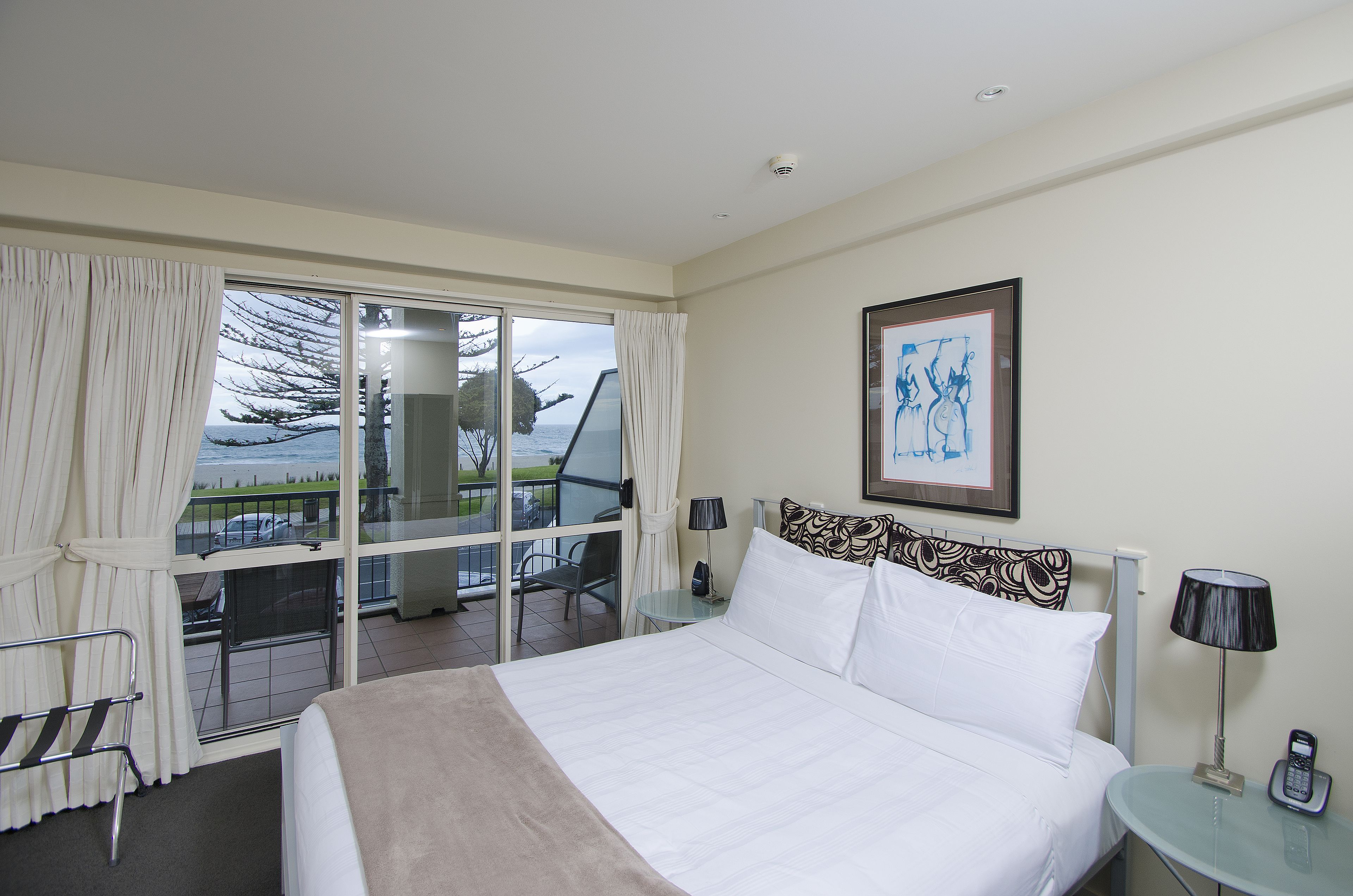 A hotel room with a bed, a chair, a table with a lamp, a phone, and a painting on the wall.