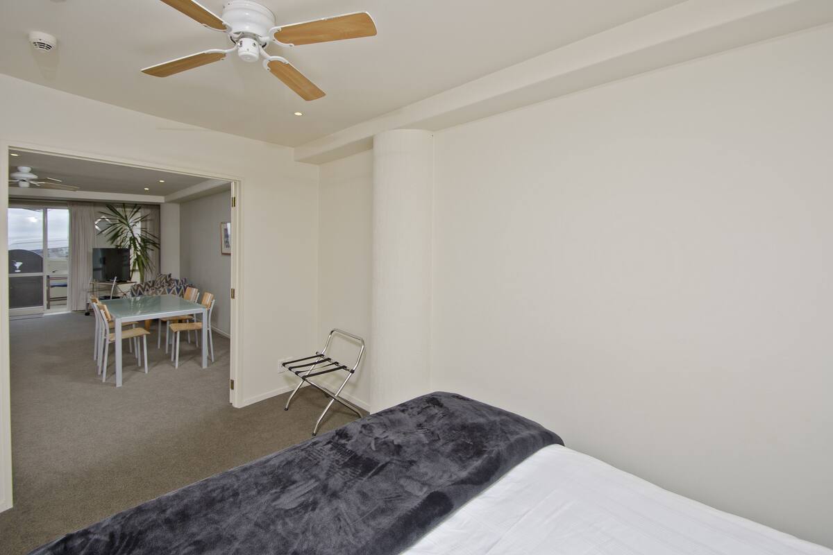 A bedroom with a bed, a ceiling fan, a dining area with a table and chairs, and a view of the outdoors.