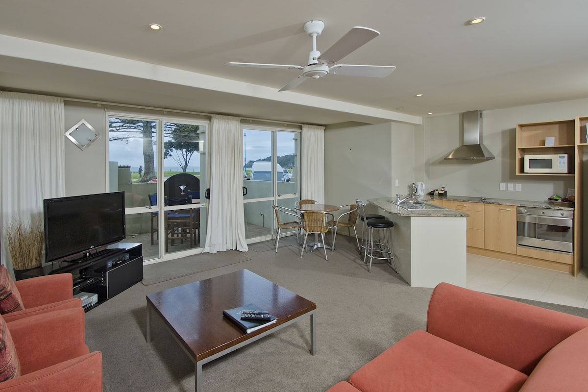 A modern living room with a flat-screen TV, a coffee table, and orange sofas. There's a dining area with a round table and chairs, and a kitchen with a microwave and stove.