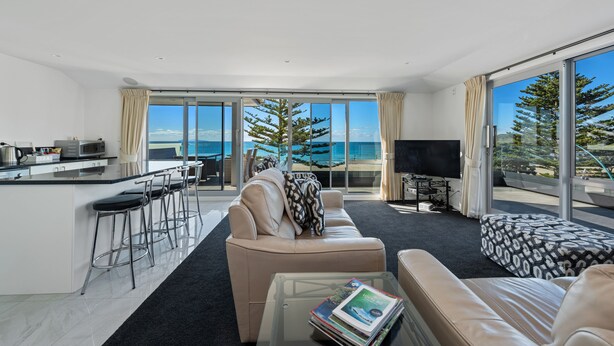 A modern living room with a sofa, a coffee table, and a TV. There is a kitchen area with a bar and stools. Large windows offer a view of the ocean and trees. - The Pavilions Beachfront Apartments (Tauranga)