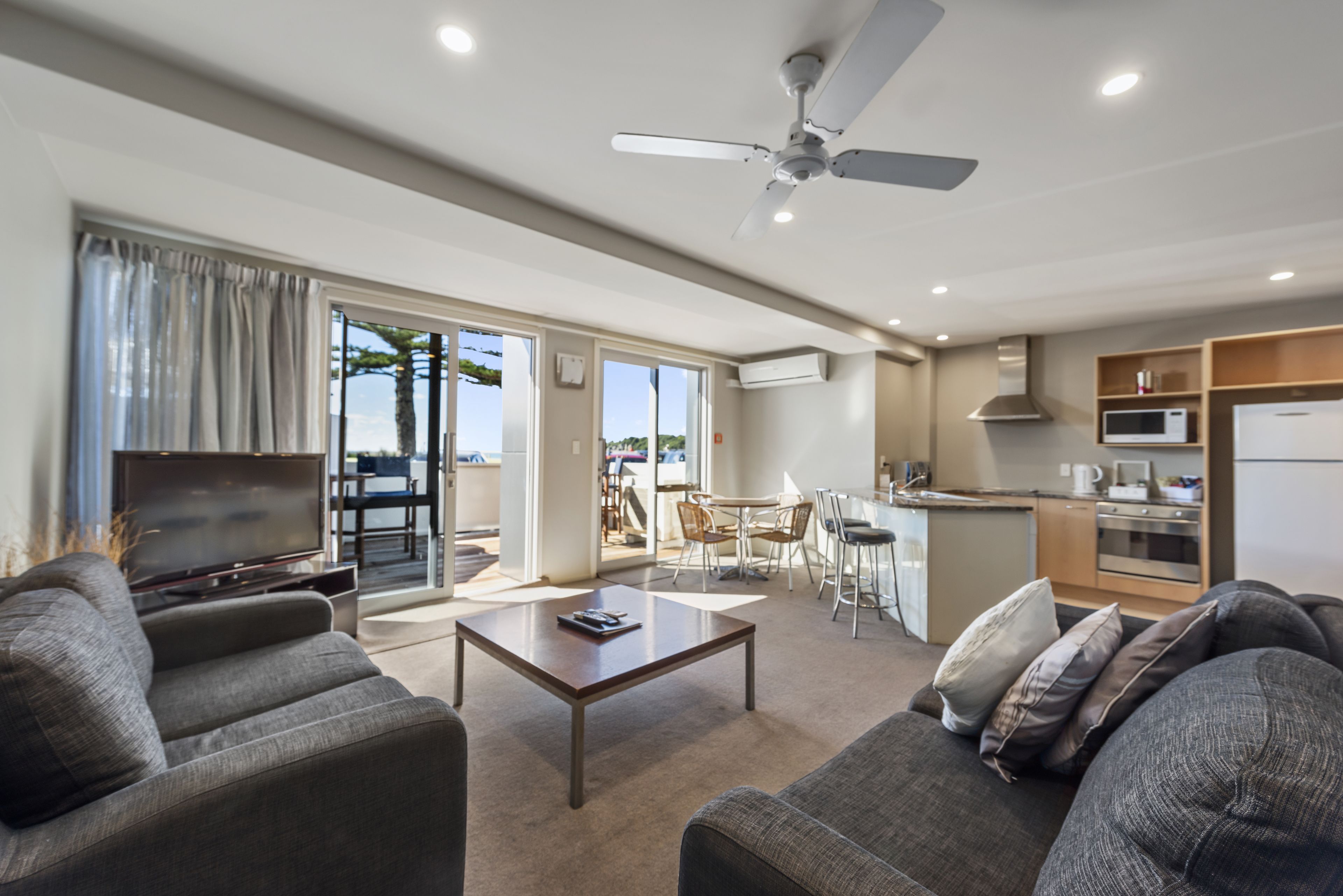 A modern living room with a flat-screen TV, a coffee table, and a sofa. There is a dining area with a table and chairs, and a kitchen area with a microwave and stove.