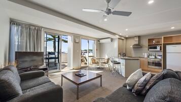 A modern living room with a flat-screen TV, a coffee table, and a sofa. There is a dining area with a table and chairs, and a kitchen area with a microwave and stove.