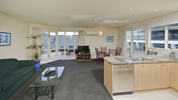 A modern apartment with a living room, kitchen, and dining area. It features a green sofa, a glass coffee table, a TV, and a potted plant. - The Pavilions Beachfront Apartments (Tauranga)