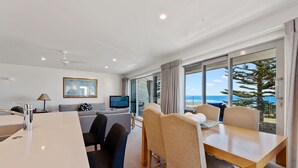 A modern hotel room with a dining area, a sofa, a TV, and a large window with a view of the ocean. - The Pavilions Beachfront Apartments (Tauranga)